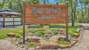 Un letrero de madera con la inscripción 'Blue Pond Retreat Montgomery, TX', rodeado por un jardín ajardinado con rocas y plantas.