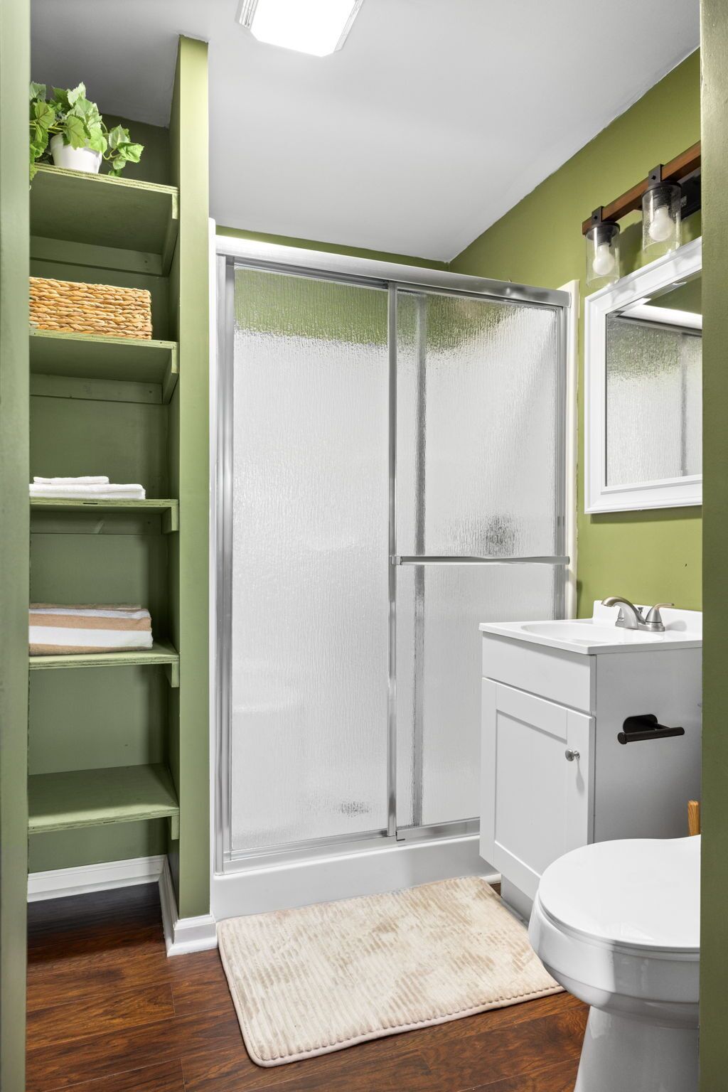 A bathroom with a glass shower door, a white sink, a mirror, and a green shelving unit.