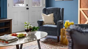 Une pièce avec un plafond en bois, un mur bleu, un fauteuil gris, une porte blanche et une grande fenêtre.