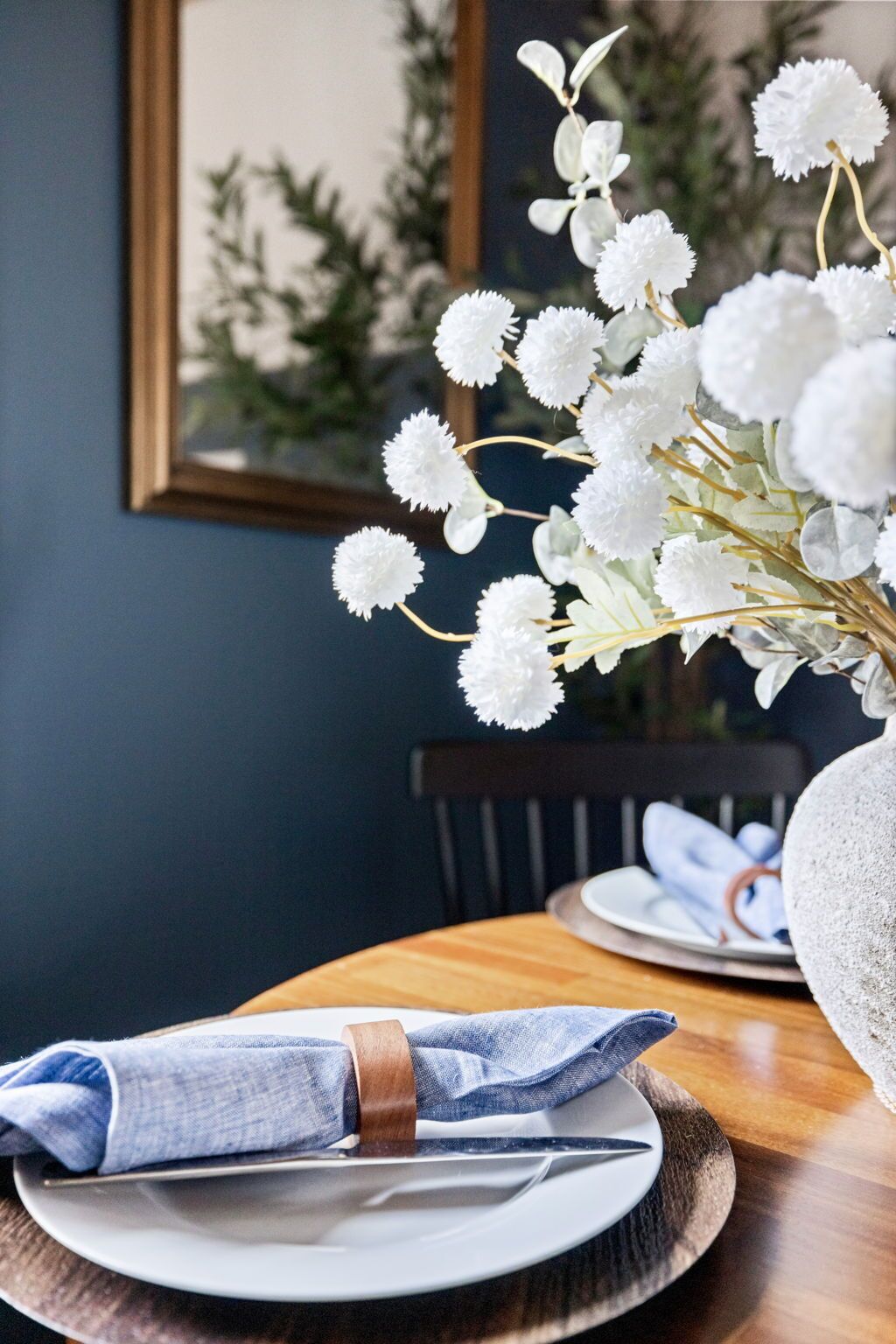 A dining table with a white plate, a folded napkin, and a knife. There is a vase with white flowers in the background.