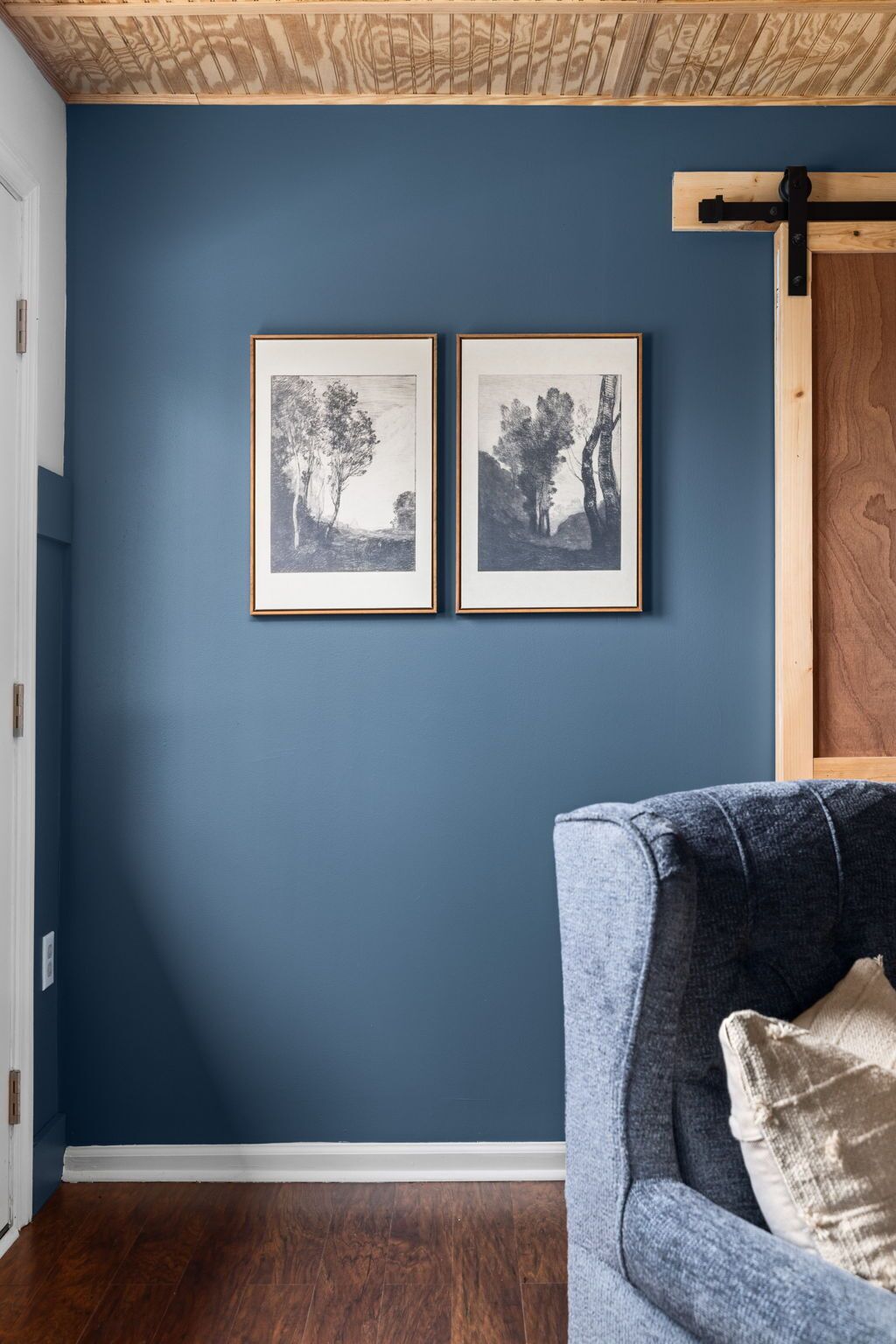 A room with dark blue walls, two framed black and white landscape photographs, a grey sofa with a beige cushion, and a wooden ceiling with exposed beams.