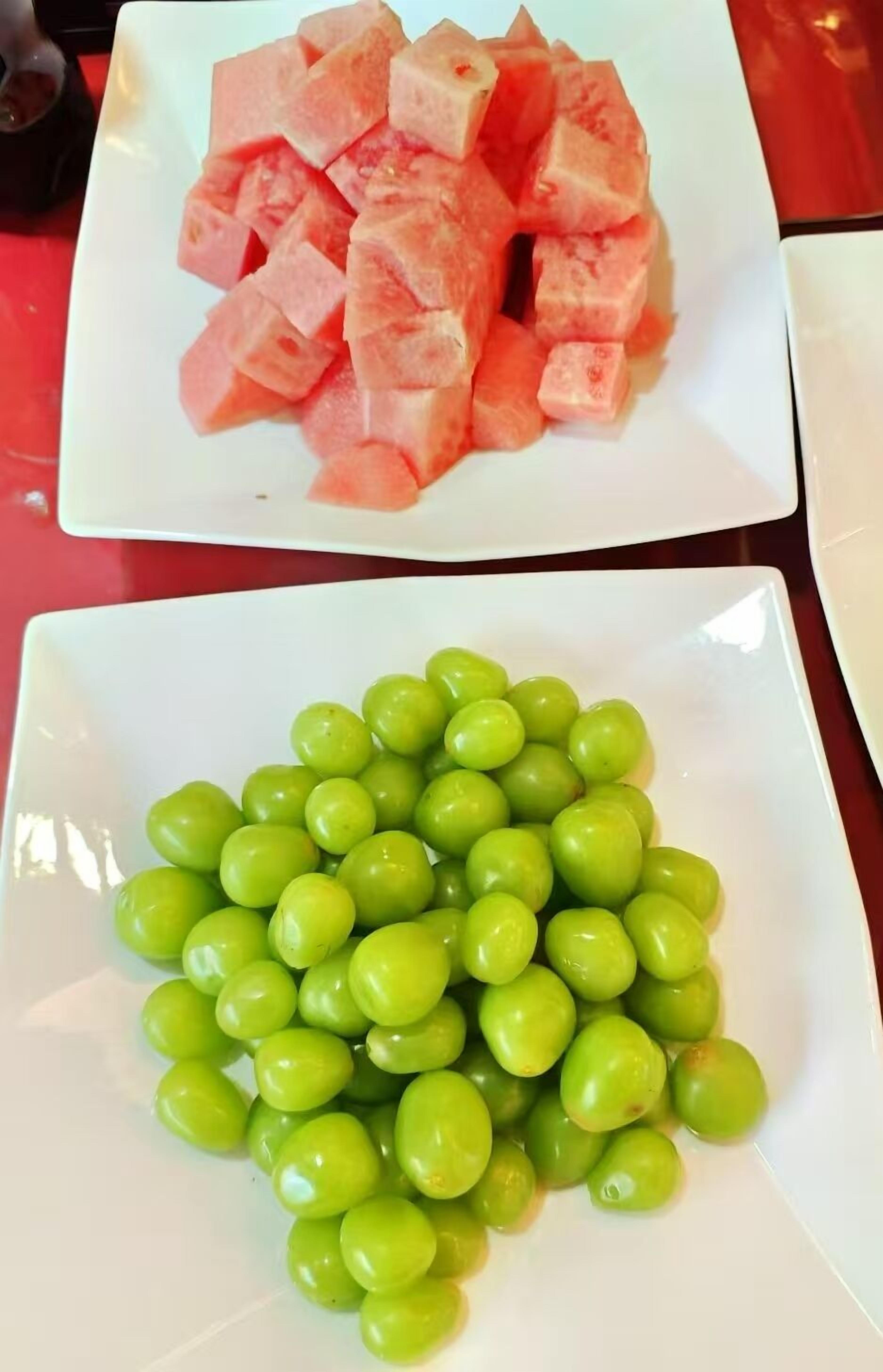 Two white plates, one with cubed watermelon and the other with green grapes.