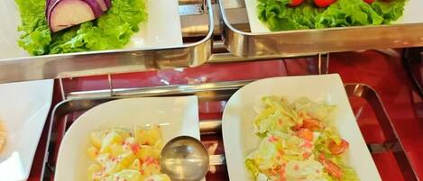 A buffet table with various dishes including sliced red onions, cherry tomatoes, pineapple chunks, and a salad with lettuce and shrimp.
