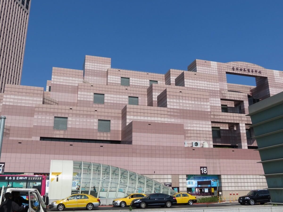 A modern building with a pink facade and a large arched entrance. There are several cars parked in front, and a sign with the number 18 is visible.