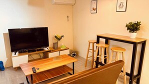 Ein modernes Wohnzimmer mit Flachbildfernseher, Holzmöbeln, einer Bank, einem kleinen Tisch mit Hocker und einer Topfpflanze.
