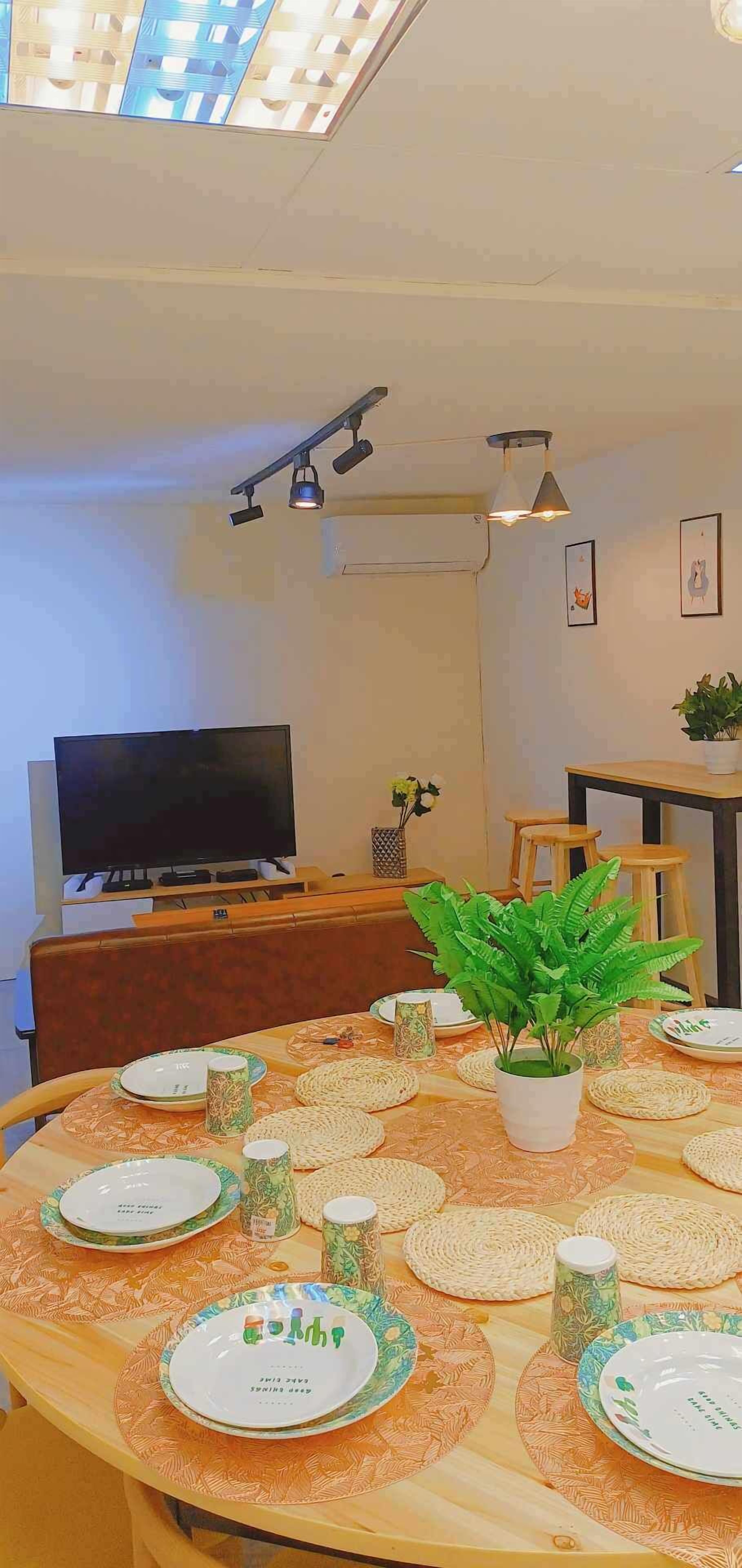 A dining area with a round table set for four, a flat-screen TV in the background, and a ceiling light fixture.