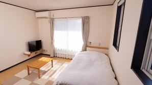 Ein kleiner, sauberer Raum mit einem Bett, einem Fernseher, einem Holztisch und einem Fenster mit Vorhängen.