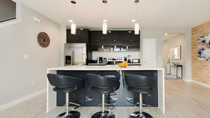 Una cocina moderna con una isla central, tres taburetes de bar, electrodomésticos de acero inoxidable e iluminación colgante.