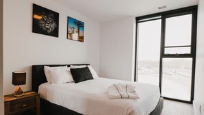 A bedroom with a large bed, a nightstand, a lamp, two wall art pieces, and a large window. - Chic Retreat w/Balcony in the Heart of Winnipeg (Winnipeg)