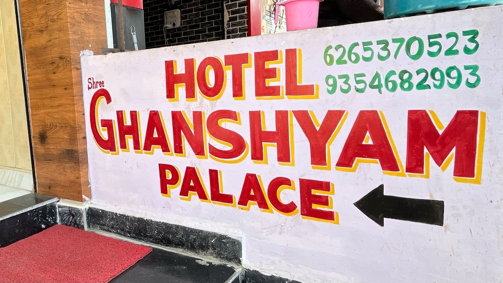 Placa do Hotel Ghanshyam Palace com números de contato e uma seta indicando a entrada.