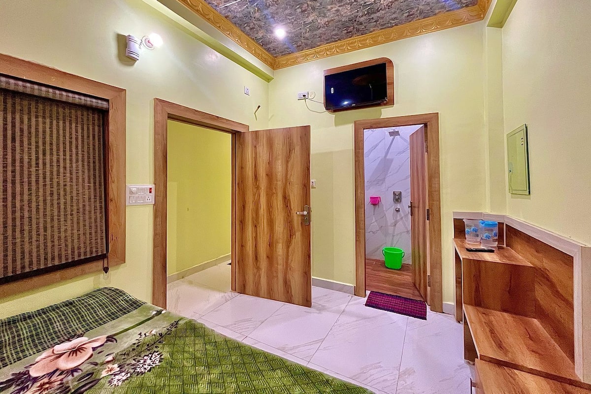 A hotel room with a wooden door, a television mounted on the wall, a green bucket, and a small shelf with toiletries.