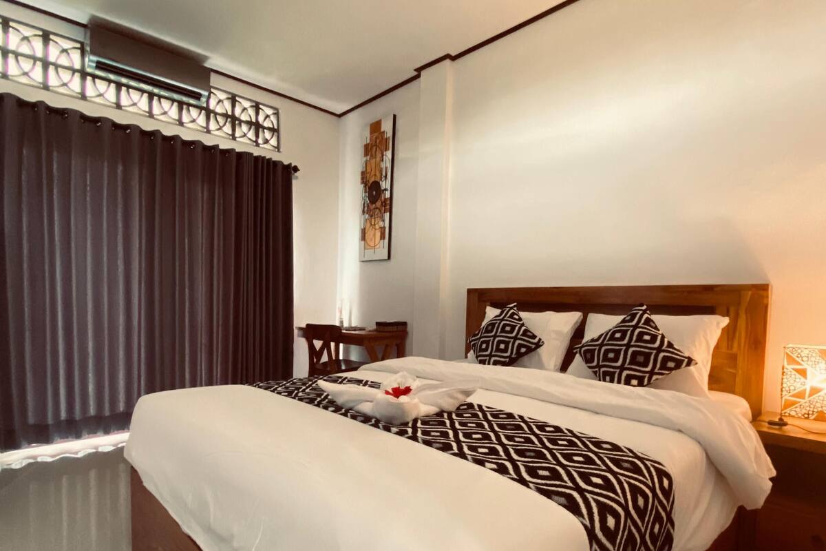 A modern hotel room with a large bed, a wooden headboard, a bedside table with a lamp, and a window with curtains.