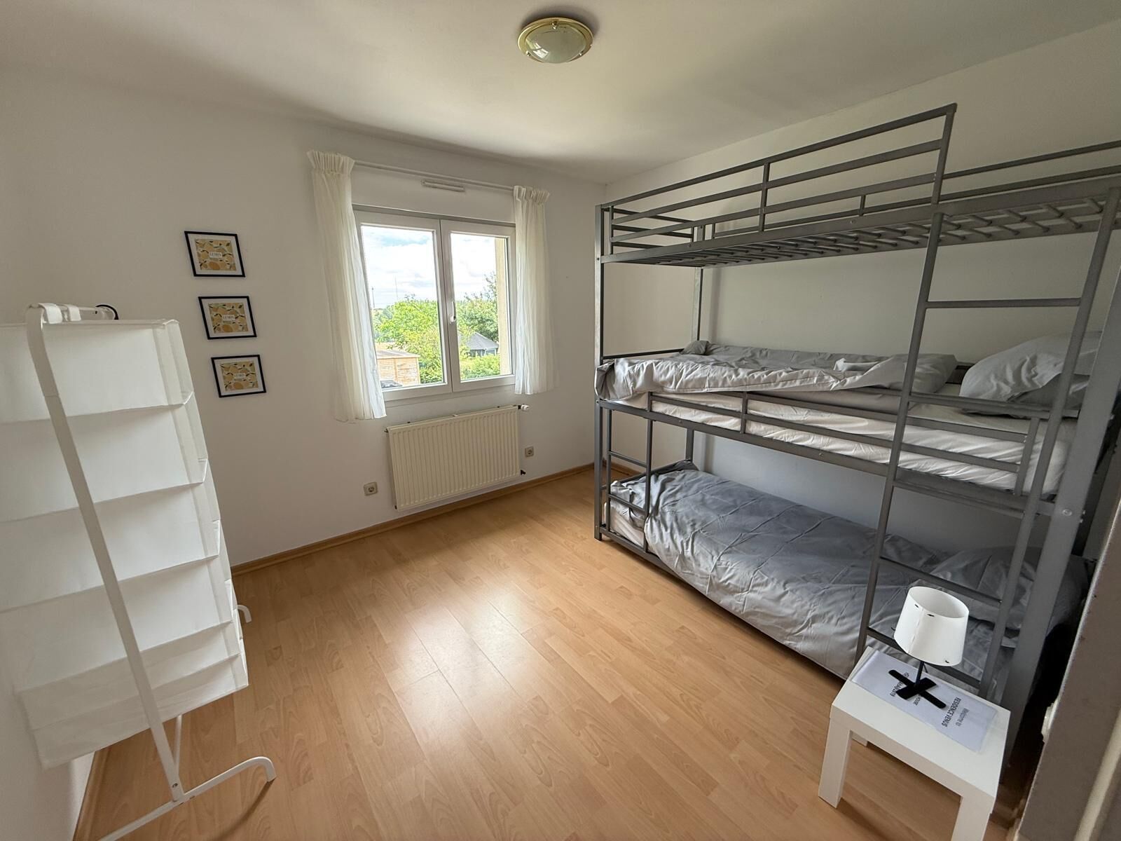 Une chambre avec un lit superposé, un radiateur blanc, une fenêtre avec des rideaux et un bureau blanc avec une lampe.