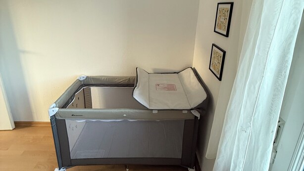 A corner room with a grey playpen, a window with white curtains, and three framed pictures on the wall.