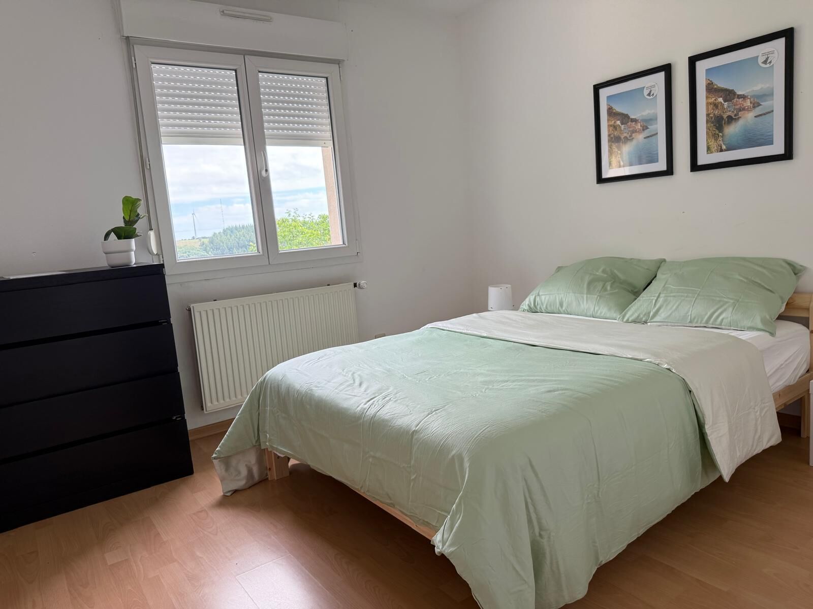 Een slaapkamer met een bed, een dressoir, een radiator, een raam met zonwering en twee ingelijste foto's aan de muur.