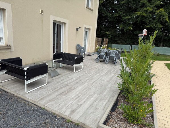 An outdoor patio with a glass-top table and two black sofas, surrounded by grey plastic chairs.
