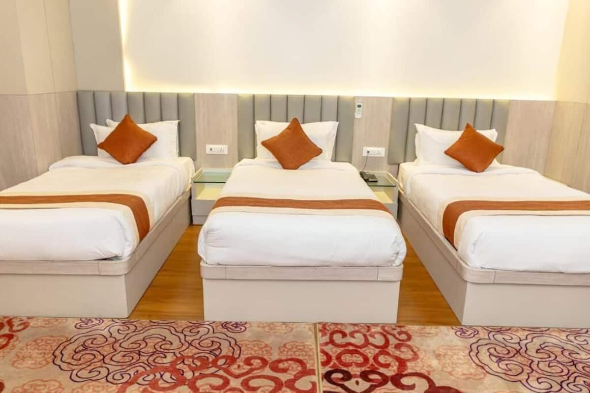 A hotel room with three beds, each with white bedding and brown pillows. The room has a modern design with a light-colored headboard and a patterned rug on the floor.