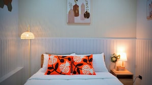 En pænt redt seng med hvide sengetøj og to orange puder med sorte mønstre, et ophængt maleri, en sengetøjslampe og et lille trænatbord med en lampe og en vase med blomster.