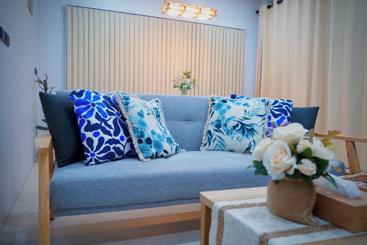 Una sala de estar moderna con un sofá gris, cojines blancos y azules estampados, una pequeña mesa de centro de madera y un jarrón con flores blancas.