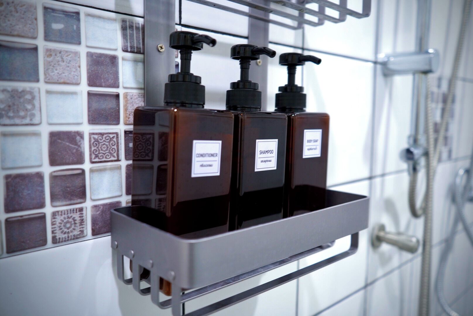 Un baño con pared de azulejos, ducha y repisa para dispensador de jabón con dos botellas que dicen "CONDITIONER" y "SHAMPOO".