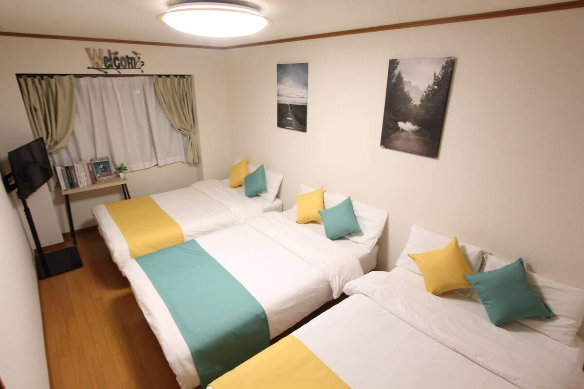 A room with two beds, each with white bedding and teal and yellow pillows. There is a desk with a chair and a TV on the left.