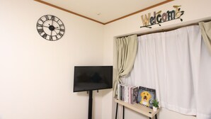 A room with a wall clock, a flat-screen TV, a 'Welcome' sign, a window with curtains, and a small shelf with books and a plant. - LUANA HOTELU NISHIKI (Tokyo)