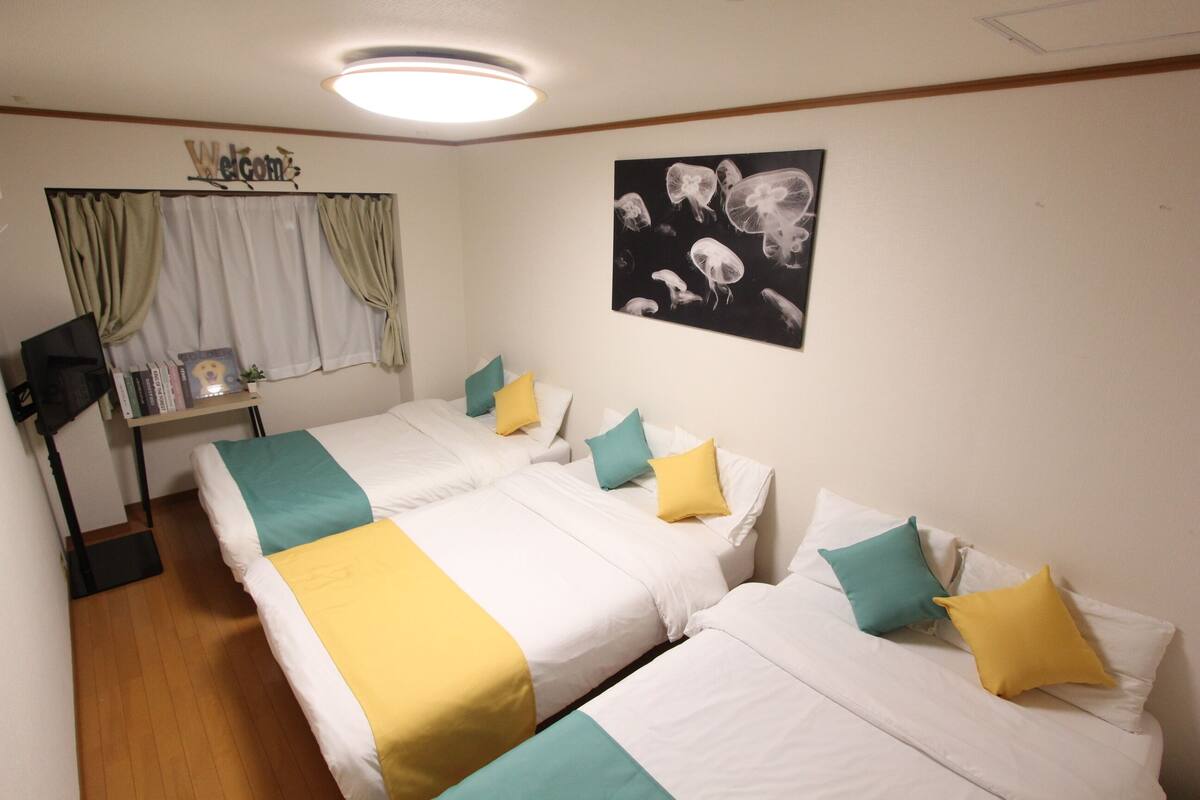 A room with two beds, each with white bedding and teal and yellow pillows. A "Welcome" sign is above the window, and a black and white jellyfish artwork is on the wall.