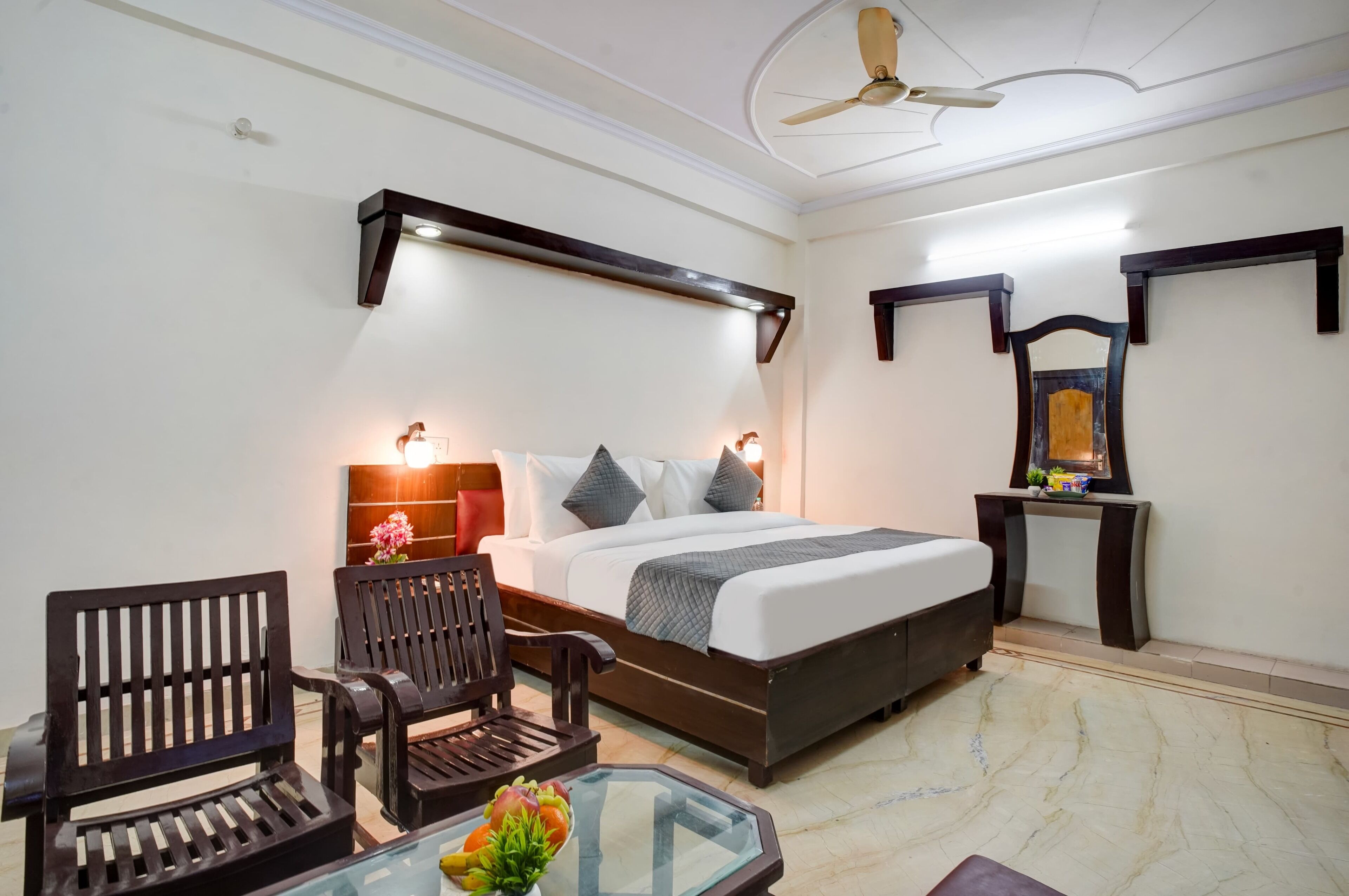 A hotel room with a bed, two wooden chairs, a glass table with a flower vase, a ceiling fan, and wall-mounted shelves.
