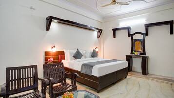 A hotel room with a bed, two wooden chairs, a glass table with a flower vase, a ceiling fan, and wall-mounted shelves.