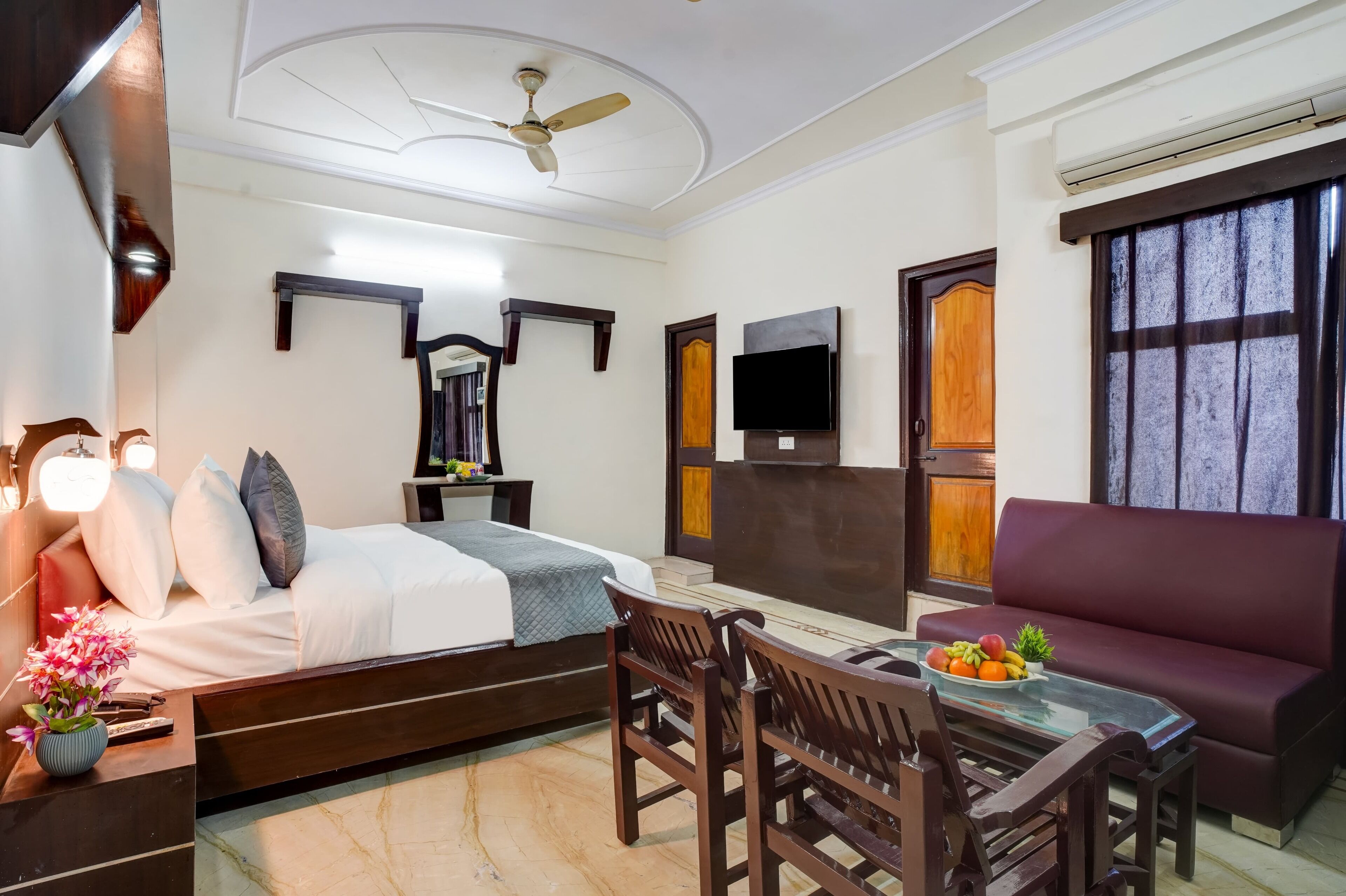 A hotel room with a bed, a TV, a ceiling fan, a sofa, a dining table with chairs, and a fruit bowl.
