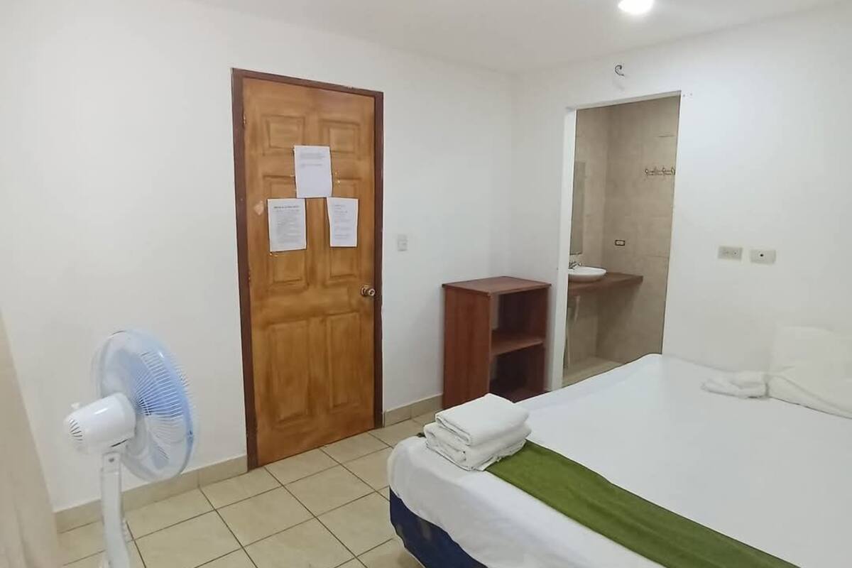 A hotel room with a bed, a wooden door with papers, a fan, and a small wooden cabinet.