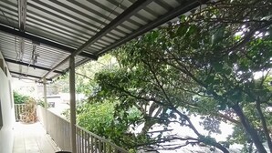 A covered balcony with a chair and a wheelbarrow, surrounded by greenery and a metal railing.