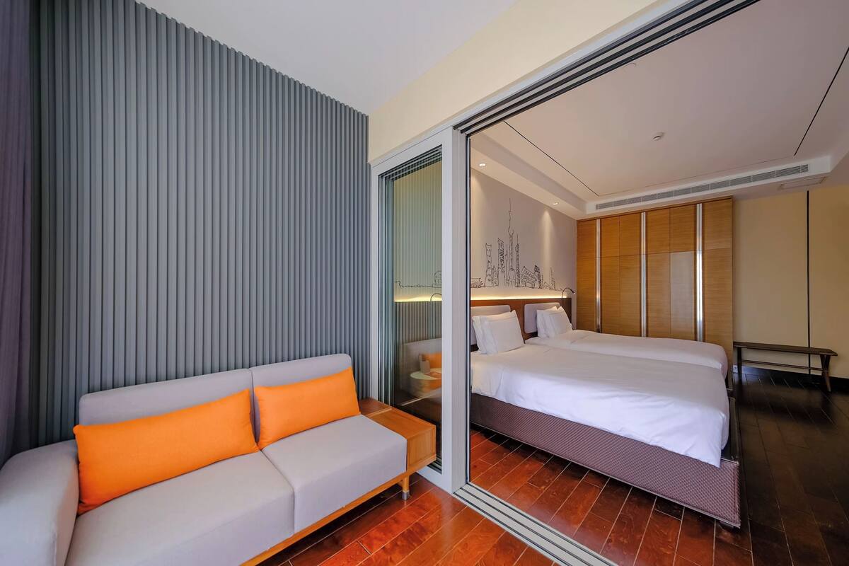 Ein modernes Hotelzimmer mit einem großen Bett, einem Sofa mit orangefarbenen Kissen und einer Spiegelwand, die das Zimmer reflektiert.