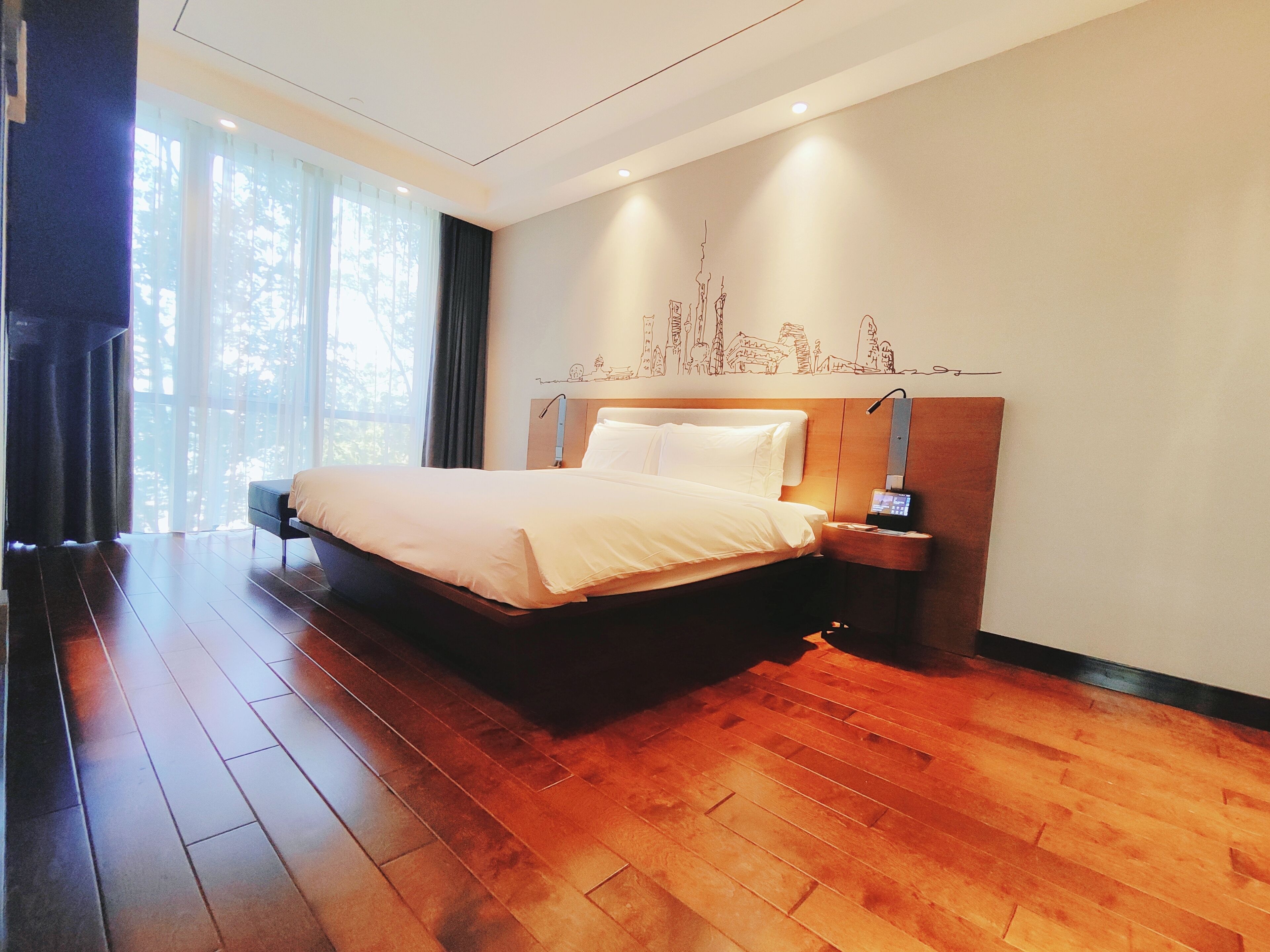 A modern hotel room with a large bed, wooden flooring, a nightstand with a lamp, and a cityscape mural on the wall.