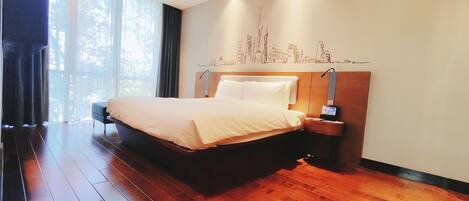 A modern hotel room with a large bed, wooden flooring, a nightstand with a lamp, and a cityscape mural on the wall.
