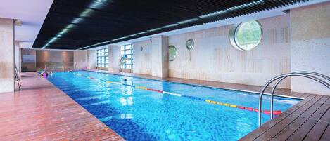 Une piscine intérieure dotée d’une terrasse en bois, de rampes en métal et de fenêtres rondes.