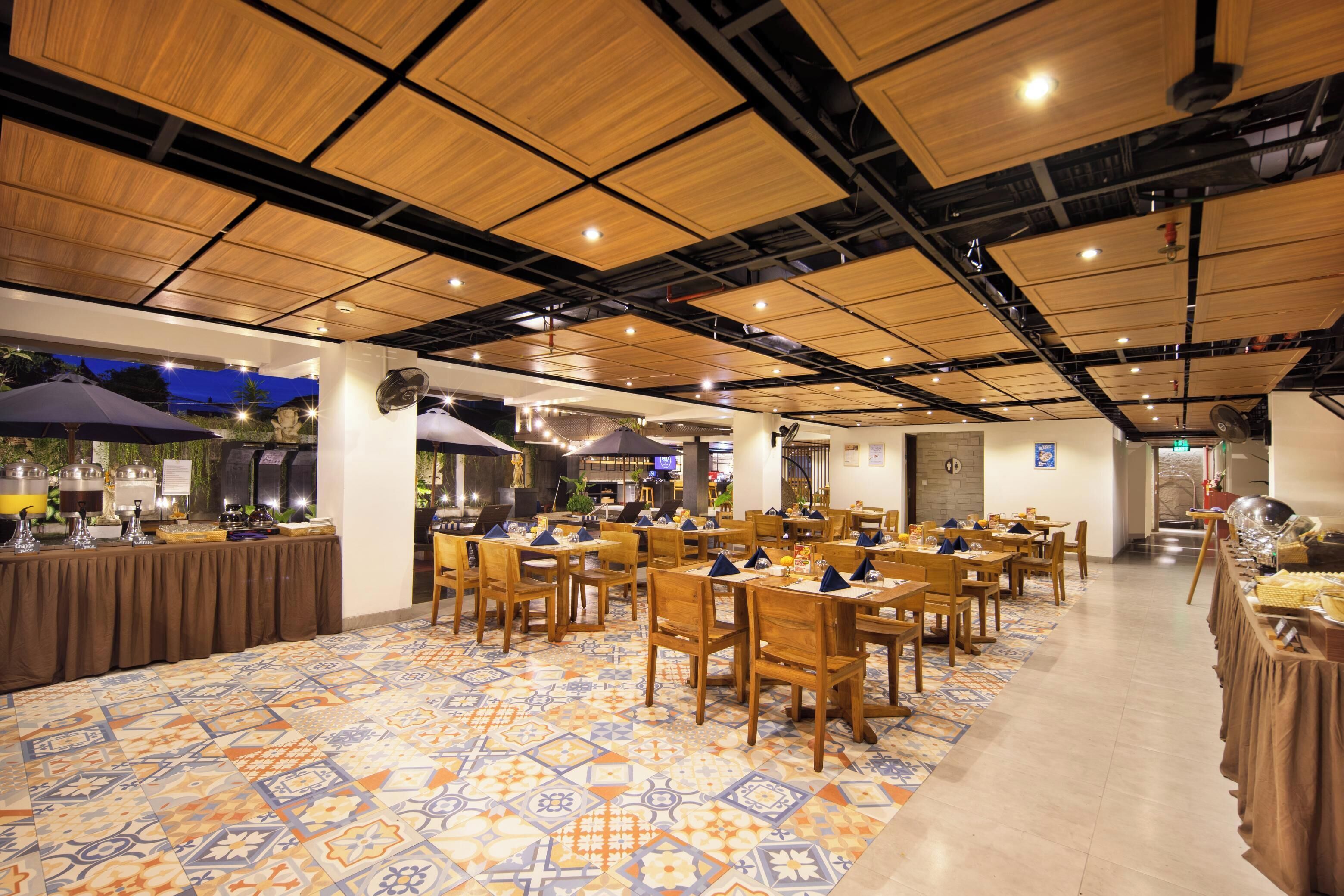 Un vaste espace intérieur aménagé en salle à manger, avec de nombreuses tables et chaises, un sol à motifs et un plafond haut avec des poutres apparentes.
