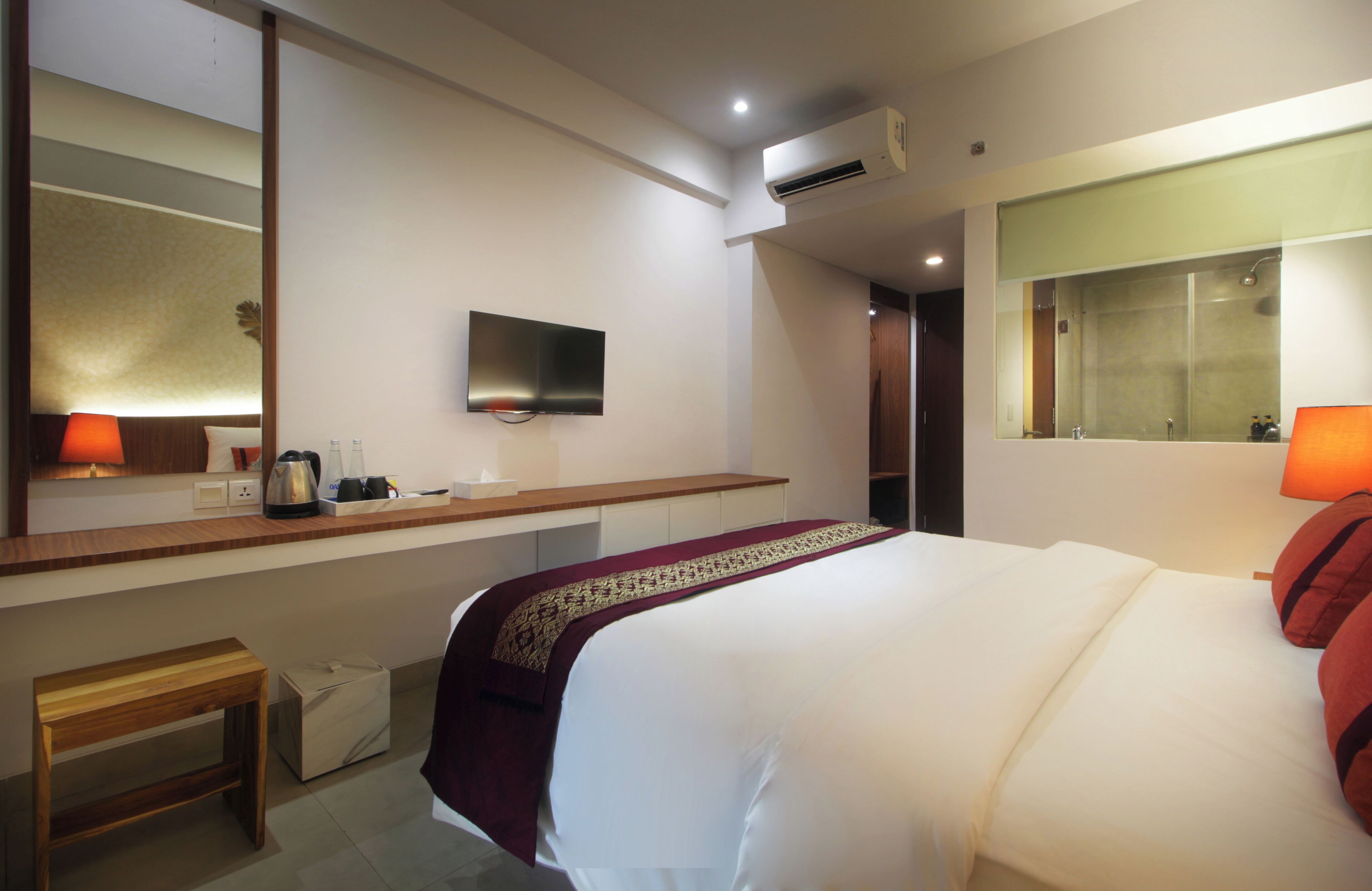 A hotel room with a large bed, a flat-screen TV mounted on the wall, a bedside table with a lamp, and a small wooden stool.