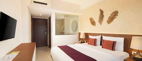 A hotel room with a bed, a desk with papers, a television, a mirror, and a wall with decorative leaves.