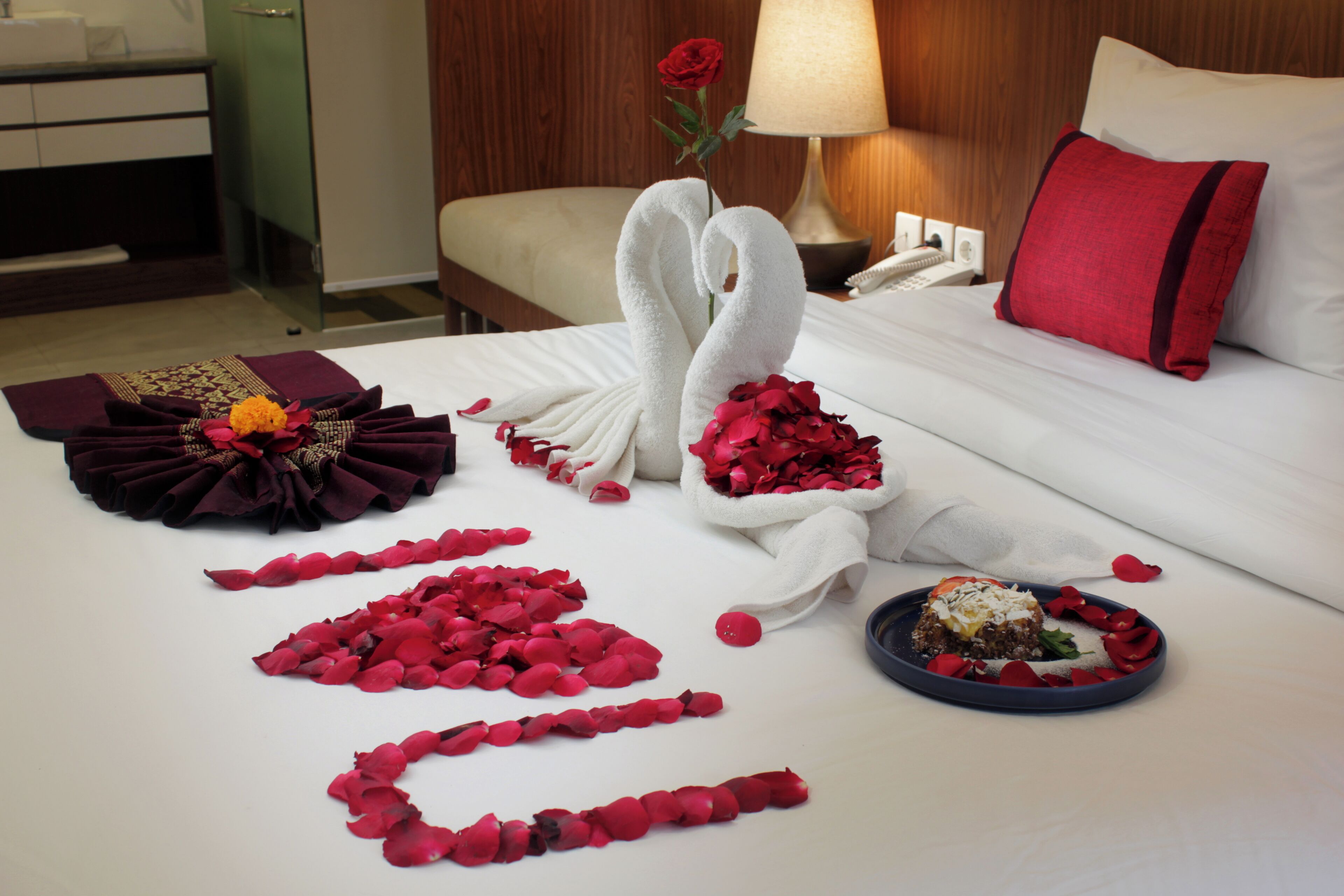 A neatly made bed with a red rose, a swan-shaped towel, and rose petals arranged on the bed.