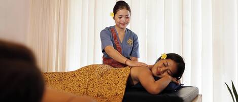 A massage therapy session in a room with a bed, curtains, and a potted plant.