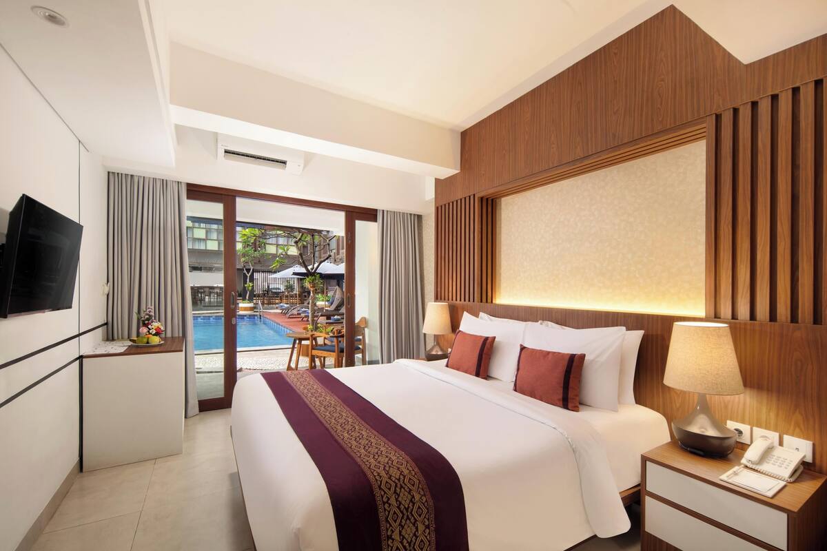 A hotel room with a bed, bedside table, lamp, TV, and a view of a pool and outdoor area.