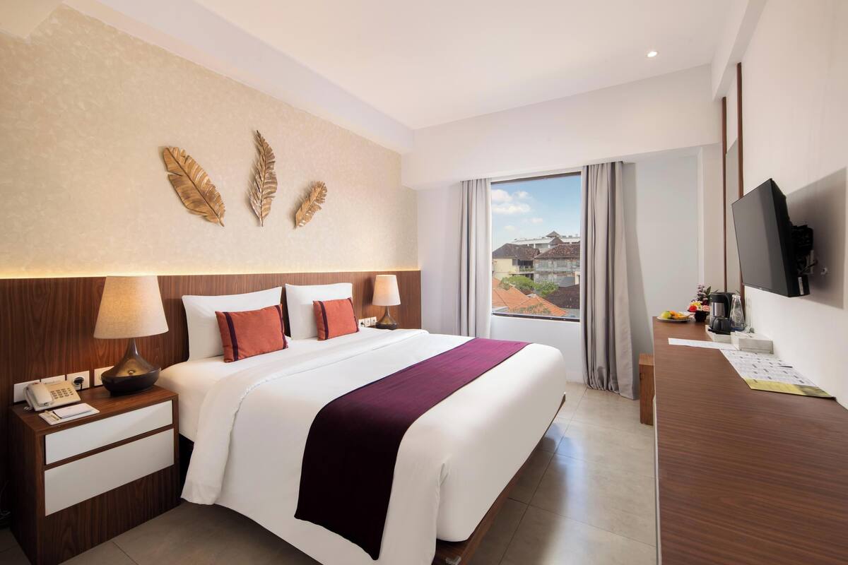 A hotel room with a large bed, a desk with a flat-screen TV, a telephone, and a window with a view of buildings.