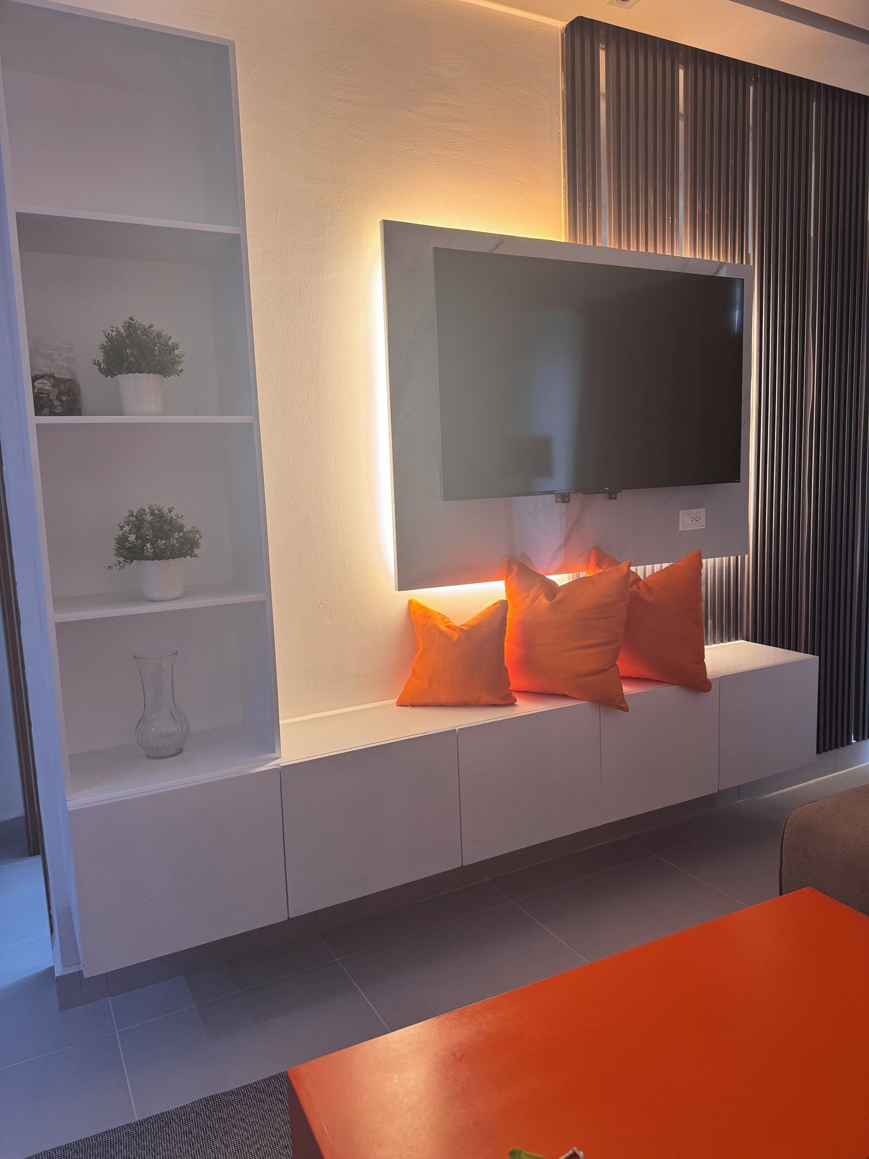 Ein modernes Wohnzimmer mit Flachbildfernseher an der Wand, einer weißen Unterhaltungseinheit mit Regalen und einem großen orangefarbenen Couchtisch.