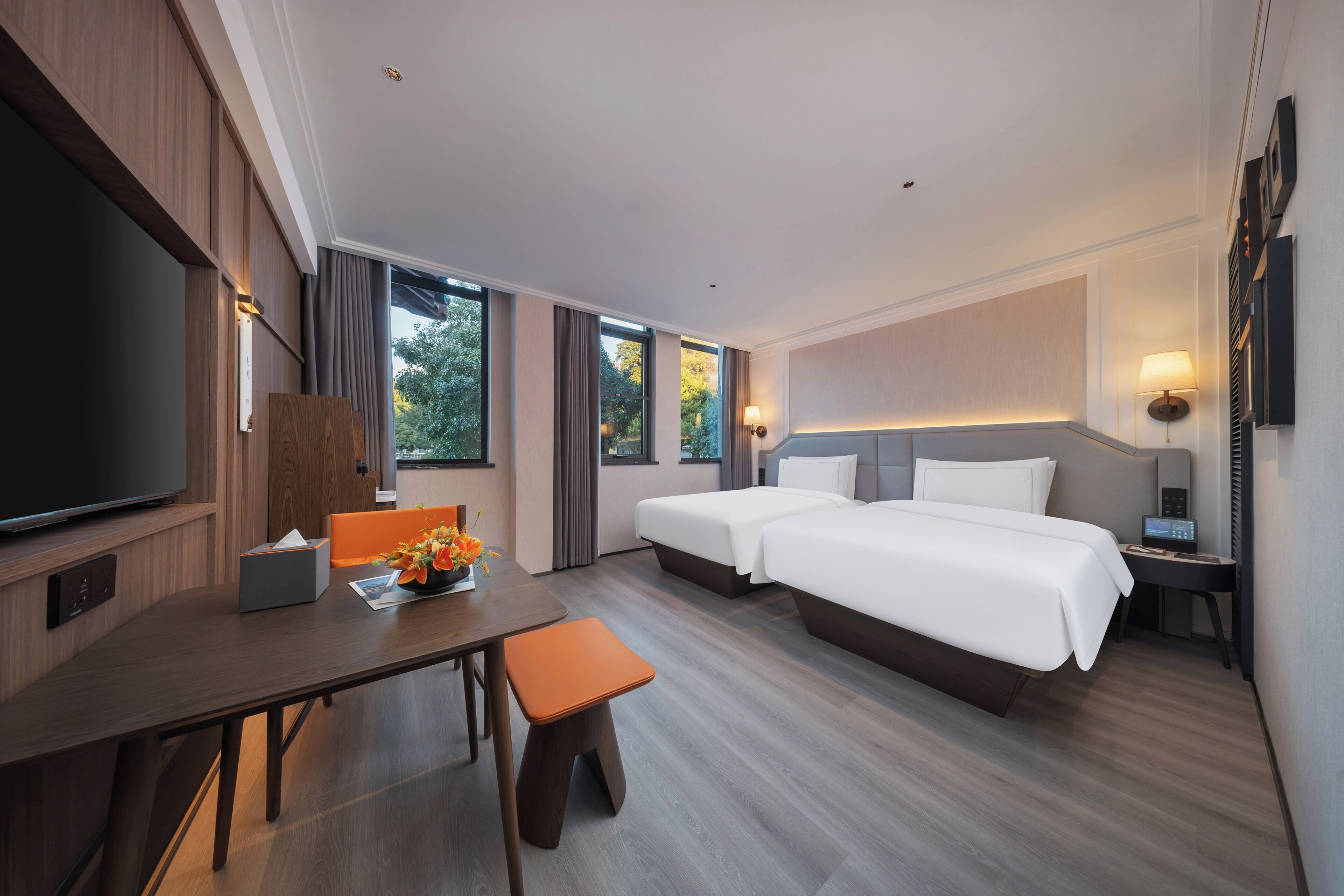 A hotel room with a large flat-screen TV, two beds, a desk with a chair, and a small table with a vase of flowers.