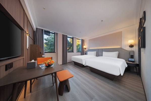 A hotel room with a large flat-screen TV, two beds, a desk with a chair, and a small table with a vase of flowers.