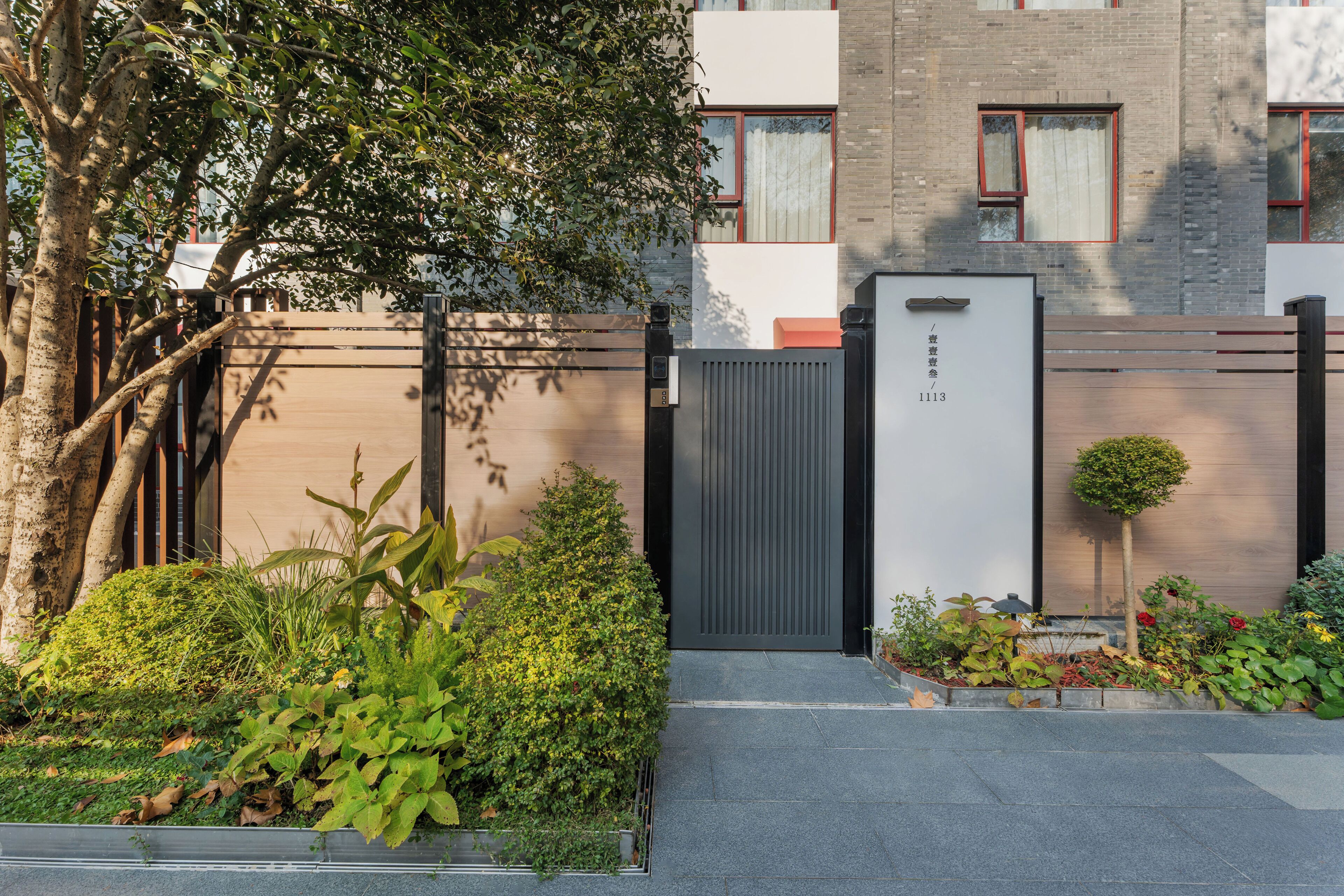 A modern building with a gated entrance, a small garden, and a tree.