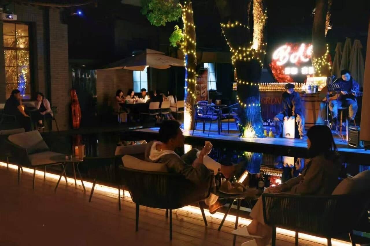 An outdoor seating area with tables and chairs, a stage with performers, and a neon sign.