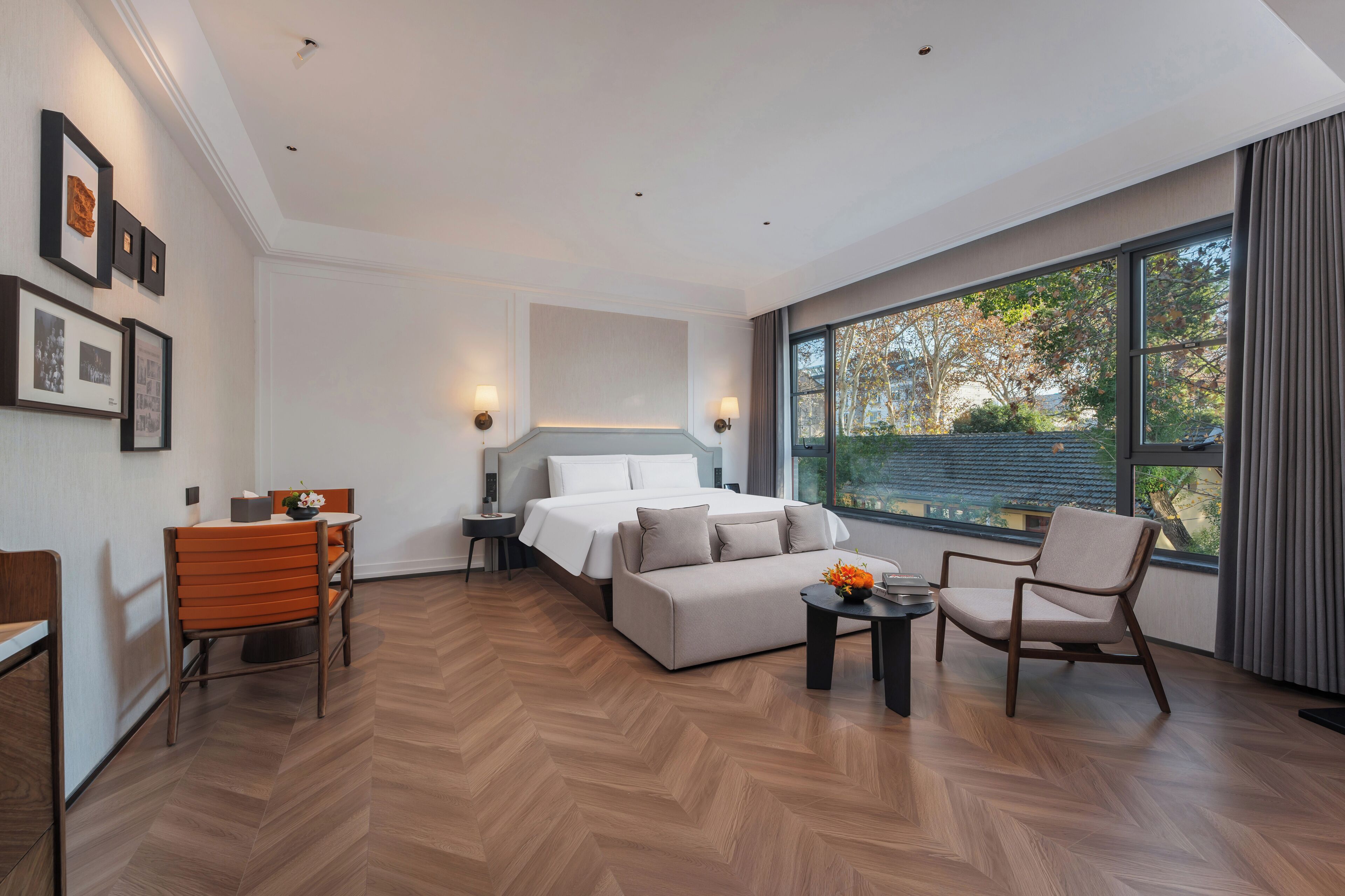 A modern hotel room with a large bed, a sofa, a dining table, and a chair. There are framed pictures on the wall and a window with a view of trees.