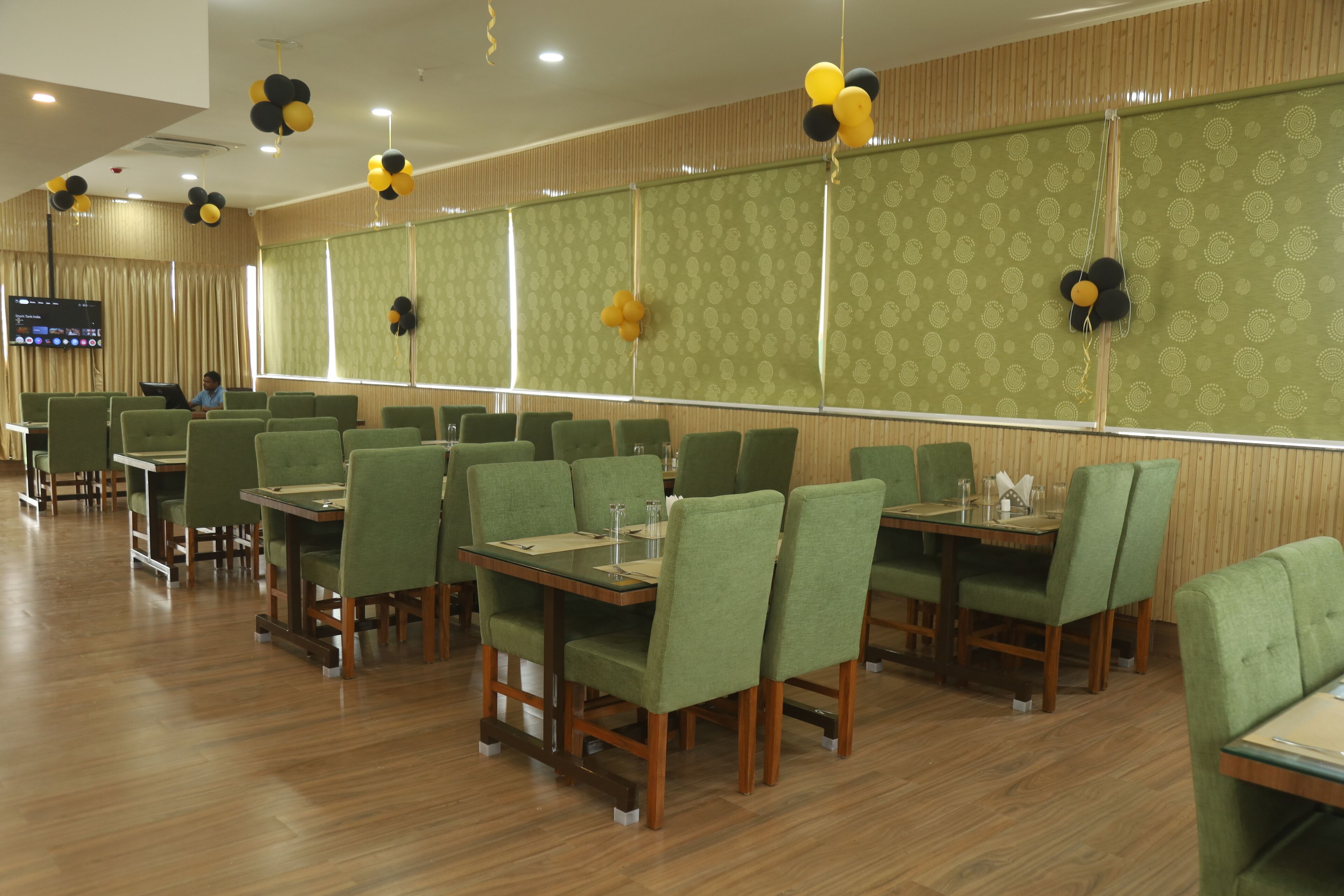 A dining area with green upholstered chairs and tables, a flat-screen TV mounted on the wall, and decorative balloons on the ceiling.